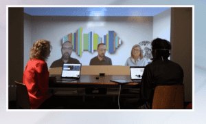 There are two people in the room on the same video call, facing a screen where there are three people sitting in a row, as though they were all sitting in the same room around a table, but three people are joining virtually