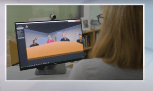 A woman is sat facing a screen with a live video call. The screen has a web camera attachment on the top. The video call looks like a virtual room where the other participants are sat across a table, as opposed to on individual window. 