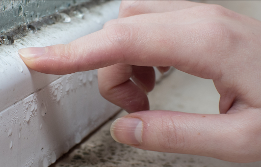 A hand touching a skirting board with signs of mould and condensation