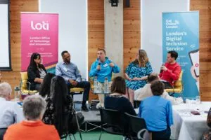 A panel of five people speaking at the front of a room at the digital inclusion conference with two banners next to them, one that states London Office of Technology and Innovation and the other banner says Get Online London