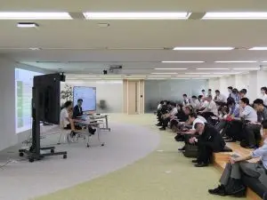 A medium outdoor horizontal shot shows approximately twenty people sitting and watching two men on a stage in a room. From left to right, a large projector screen features a presentation slide, partially in focus, that is visible on a large wall and a flat TV stand with another flat TV next to it. Below the flat TV is a beige table where two men with laptops are sitting, both men wear white shirts and have black hair. One man is sitting on a white chair, and the other is sitting on a white and green chair. Both of them face a big audience sitting on some steps of an auditorium. The steps are beige wood with some dark blue and black cushions. Behind the seating area, an elongated room features a white wall and a white wooden door in the center and some windows next to the door. Approximately, fifteen people are sitting on the steps with different dark-colored clothes, most of them wearing white shirts and dark pants and shoes. Some people are holding laptops on their laps. Some people are facing the stage and some are facing down. A beige circular carpet can be found from left to right covering half the area from the stage to the seating area. Another green carpet surrounds the other part of the room. Overhead lightings are turned on, making the room bright. Multiple electrical wires can be found coming from the table towards the large wall. A projection lamp is visible coming from the ceiling, close to the center of the image. The picture quality is slightly grainy.