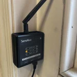 A photograph of a sensor attached to a wall by a window, the sensor is shaped like a black box with an antenna
