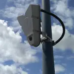 A photo of a sensor attached to a lamp post with the sky in the background. The sensor is shaped like an inverse triangle.