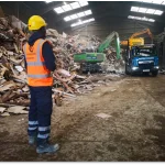 Image of a man wearing high visability clothing and a hard hat standing in a big hanger with industrial waster and machinary that is moving the waste